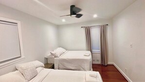 Quarto com duas camas, ventilador de teto, janela com cortinas e parede branca.