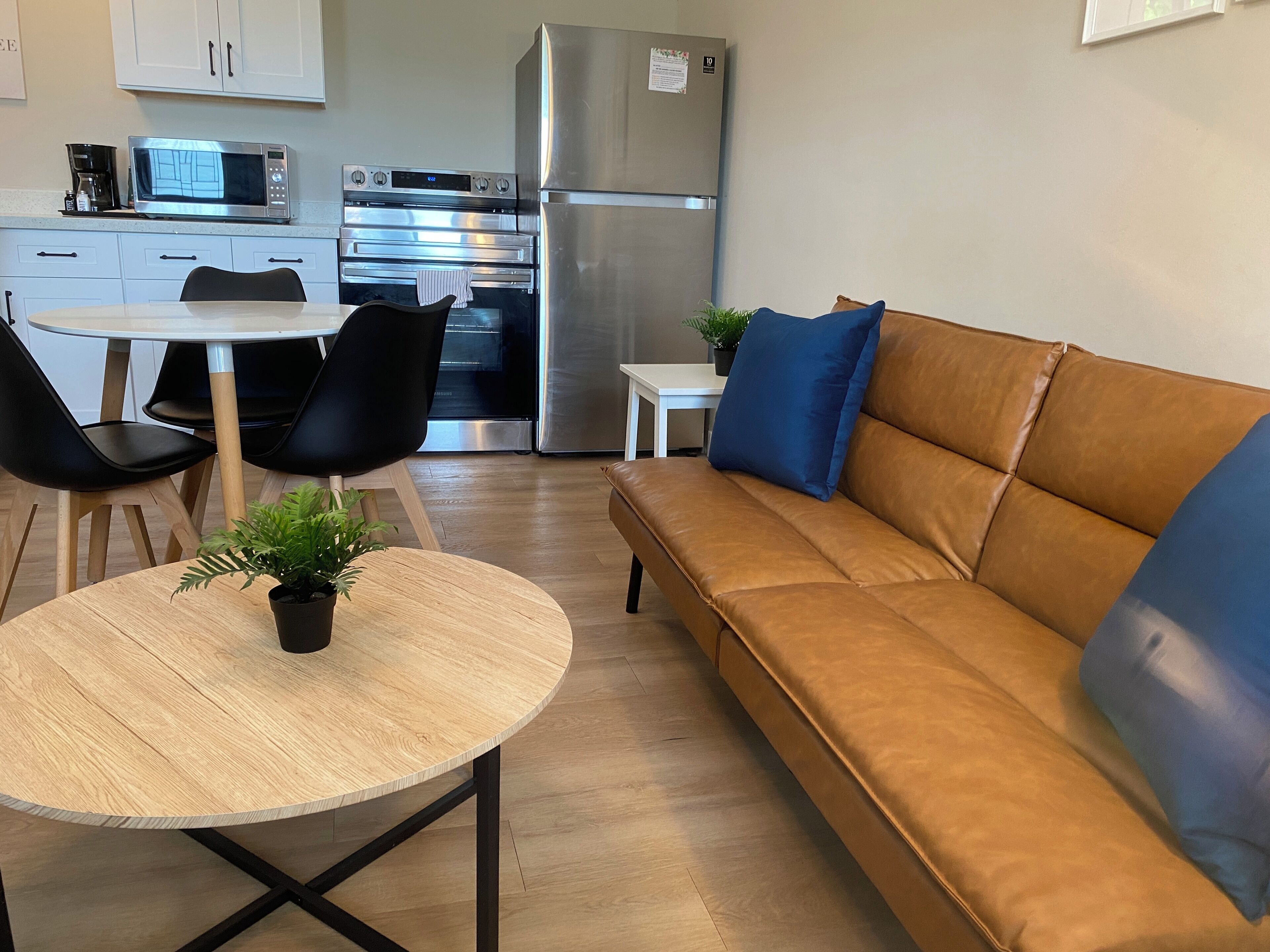 A modern living room with a brown leather sofa, a round wooden coffee table, and black chairs. A small white table with a potted plant is in the background.