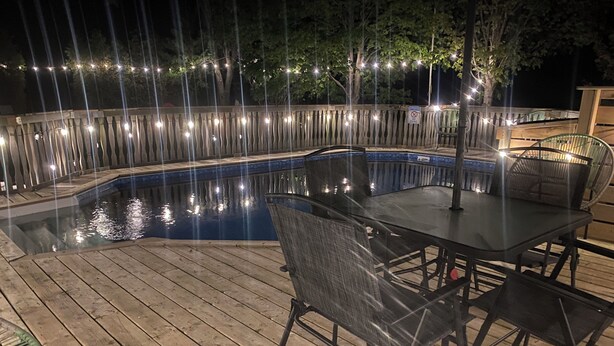 A wooden deck with a glass-top table and chairs, surrounded by a pool and string lights. - South Shore Suites (Bayfield)