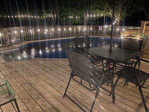 Ett trädäck med ett bord och stolar med glasskiva som bordsskiva, omgivet av en pool och ljusslingor.