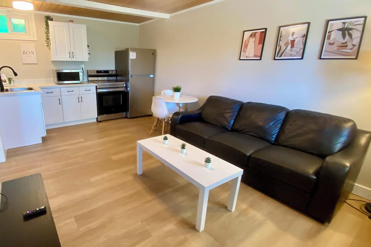A modern living room with a black leather sofa, a white coffee table with plants, and a kitchen in the background.