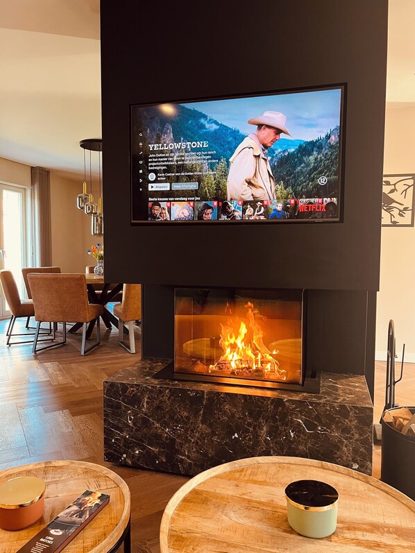 A modern living room with a flat-screen TV mounted above a fireplace, a dining area with chairs, and a wooden coffee table.