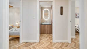 Ein modernes Schlafzimmer mit Holzboden im Fischgrätmuster, einem Bett mit grauer Steudecke, einem Nachttisch mit Lampe und Blick in ein Badezimmer mit Spiegel und Waschbecken.