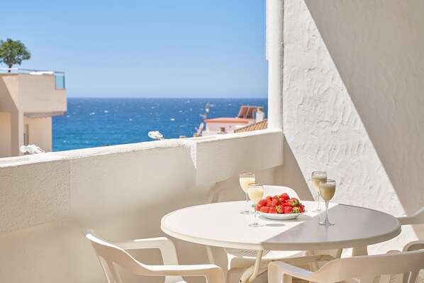 A round white table with two chairs, a bowl of strawberries, and two glasses of white wine on a balcony overlooking the sea.