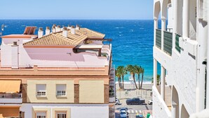 Ein Küstengebäude mit Terrasse, das auf das Meer blickt, eine Straße mit parkenden Autos und Palmen.