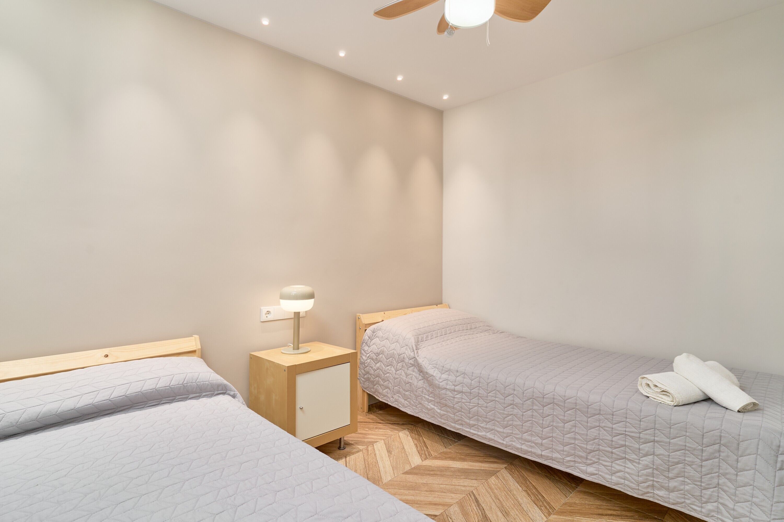 A bedroom with two beds, a nightstand with a lamp, and a ceiling fan.