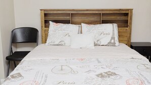Een bed met een dekbed en kussens met Parijs thema, een houten hoofdbord, een zwarte stoel en een klein nachtkastje.