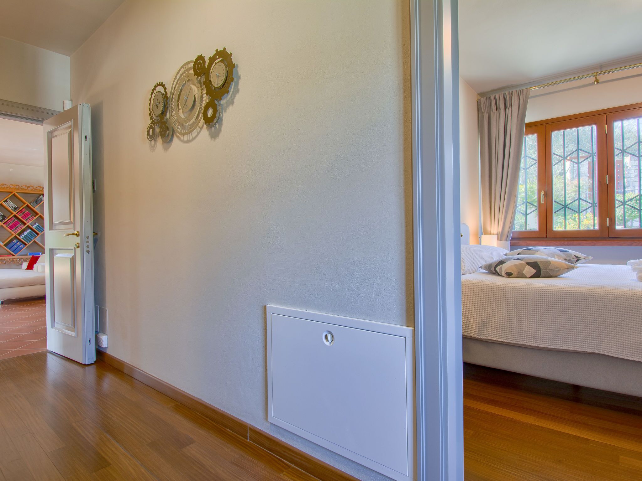 Una camera da letto con un letto, una finestra con tende, una sveglia a muro e una porta che dà su un'altra stanza.