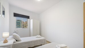 A hotel room with two beds, a bedside table with a lamp, a window with a view of buildings, and a door. - Vacation 'Ifae 3' with Wi-Fi (Guía de Isora)