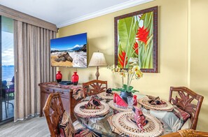 A dining area with a glass table, woven placemats, and chairs. There is a painting of a beach scene and a tropical flower on the wall.
