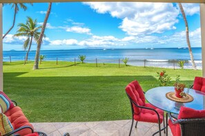 A patio with a glass table and red chairs, overlooking a grassy area with palm trees and a view of the ocean.