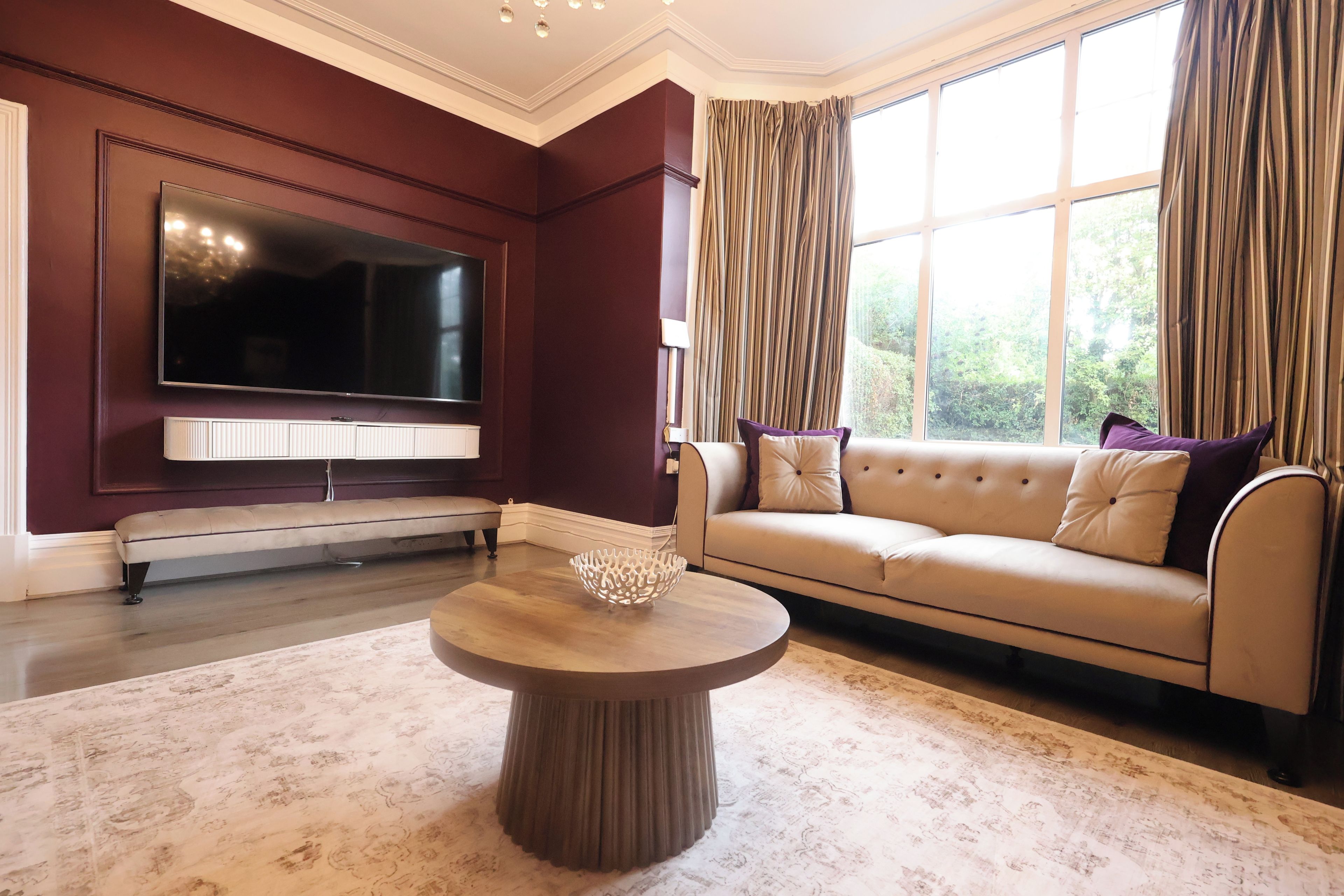 Un salon doté d’un grand téléviseur à écran plat fixé au mur, d’un canapé beige agrémenté de coussins, d’une table basse ronde et d’un tapis à motifs.