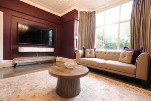 A living room with a large flat-screen TV mounted on the wall, a beige sofa with cushions, a round coffee table, and a patterned rug.