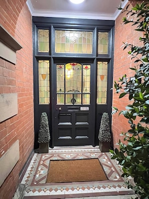 A black door with a sign instructing to use the post box on the wall, flanked by two decorative urns, leading to a tiled entrance.