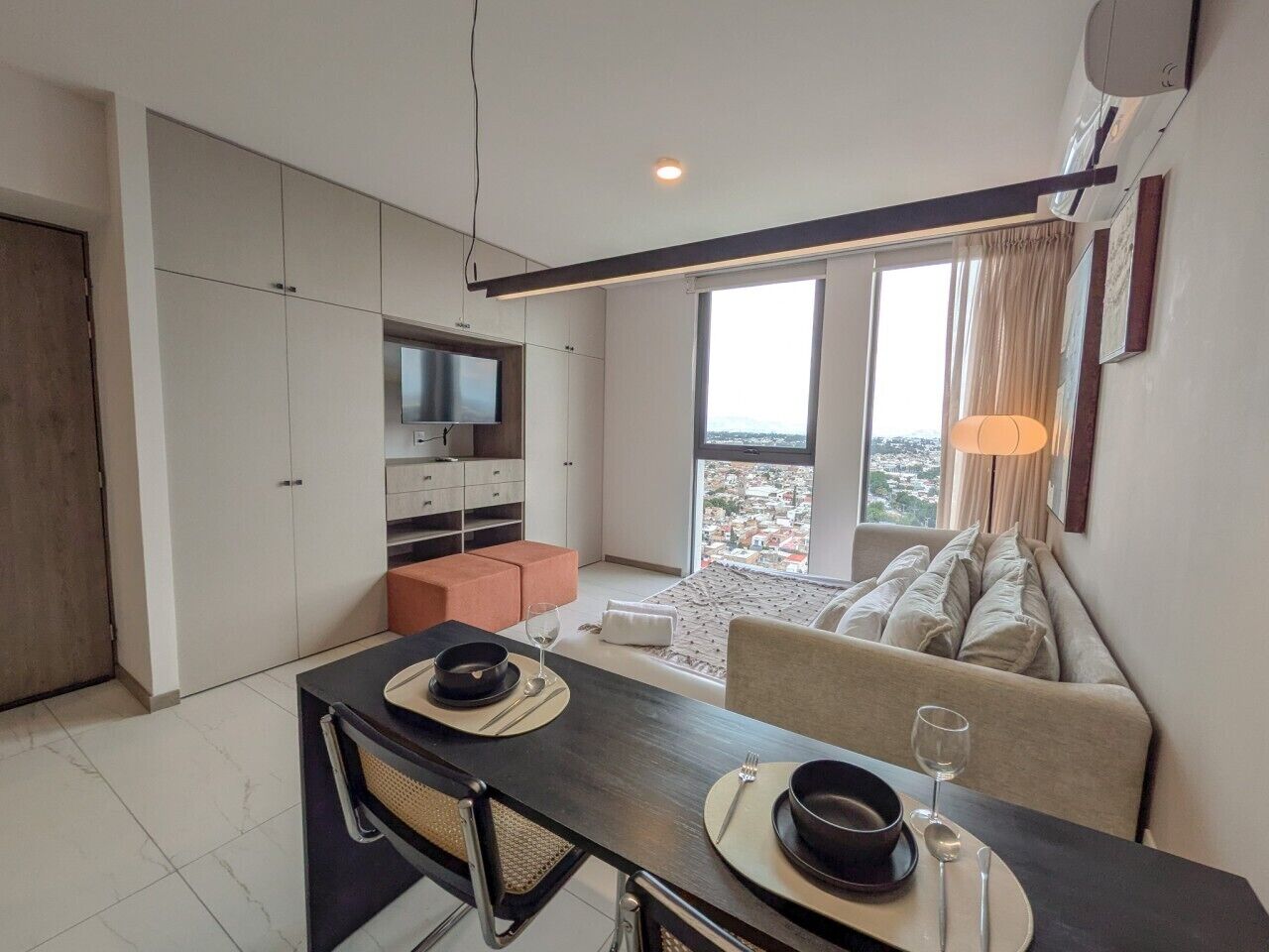 A modern living space with a dining area, a sofa, a television, and a view of the city.