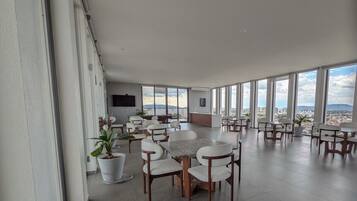 A spacious dining area with multiple tables and chairs, large windows offering a view of the city, and a flat-screen TV mounted on the wall.