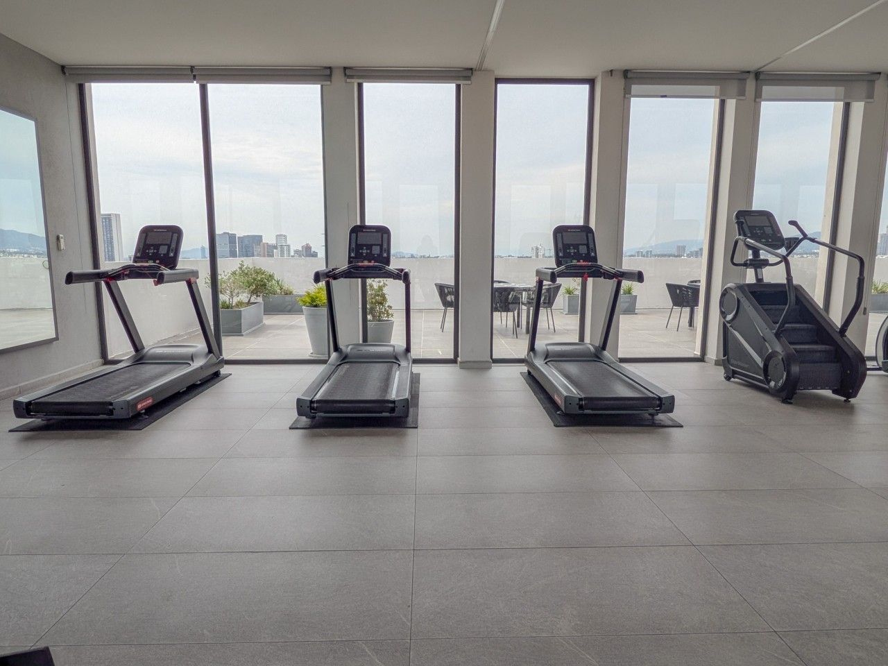 A gym with treadmills and an escalator, large windows, and a view of a cityscape.
