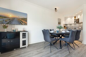 A modern dining area with a wooden table set for six, surrounded by dark chairs. A large painting of a sunset over a beach is on the wall. - An der Seedüne, App. Carpe diem - "Relax in our vacation apartment at (Unknown)