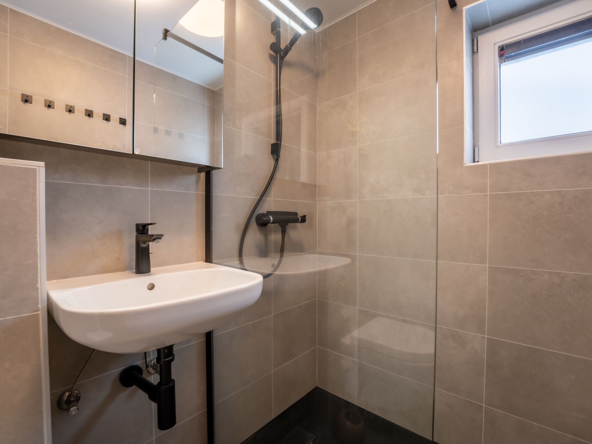 Un baño moderno con un lavamanos blanco, un cabezal de ducha de mano y una ventana sobre el lavamanos.
