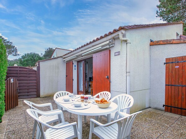 Een terras met een tafel voor vier personen, omringd door witte stoelen, voor een gebouw met een rode deur en een bord met het nummer 175.