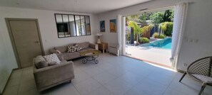 Una sala de estar con sofá, mesa de centro, una silla, televisor, espejo y puerta corrediza de vidrio que da a una piscina.