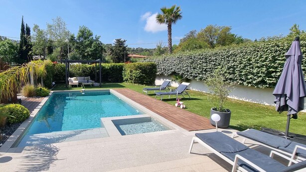 A rectangular swimming pool with a wooden deck, surrounded by a well-maintained garden with various plants and a pergola.