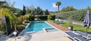 Une piscine rectangulaire dotée d’une terrasse en bois, entourée d’un jardin bien entretenu où poussent différentes espèces de plantes, et agrémentée d’une pergola.
