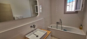 Une salle de bain avec un lavabo blanc, une baignoire, un miroir et une fenêtre.