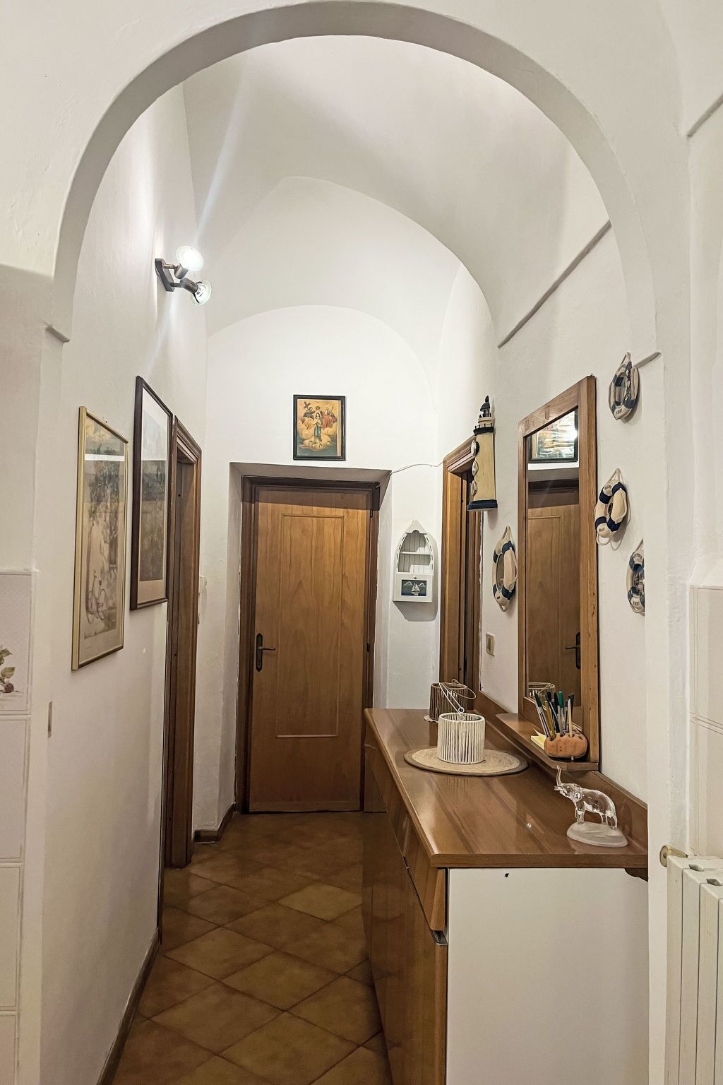 A hallway with a wooden door, a white wall with framed pictures, a wooden counter with a sink, and a tiled floor.