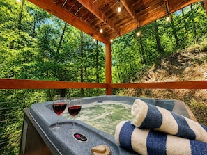 A hot tub with two glasses of red wine, a towel, and a digital control panel, set against a forest backdrop. - Cove Creek Overlook – Creek, Hot Tub & Fire Pit (Sevierville)