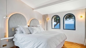 Ein Schlafzimmer mit einem großen Bett, zwei Rundbogenfenstern mit Meerblick, einer runden Wandleuchte und einem Nachttisch mit Lampe.