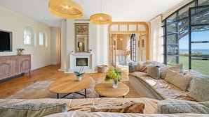 Ein geräumiges Wohnzimmer mit einer großen Couch, einem Holztisch und einem Kamin. Große Fenster bieten einen Blick ins Freie.