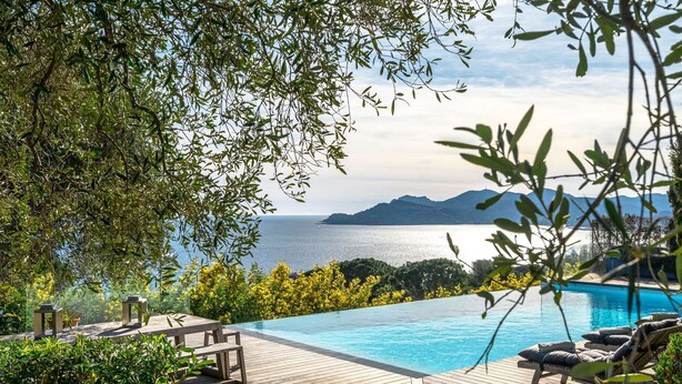 Ein Pool ohne Ende mit einer Holzterrasse, Liegestühlen und Blick auf das Meer und die Berge.