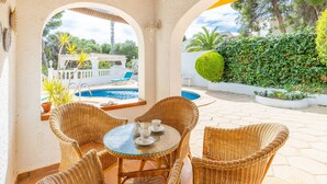 A covered outdoor seating area with wicker furniture and a glass table, overlooking a swimming pool and lush greenery. - Carlota by Interhome (Moraira)