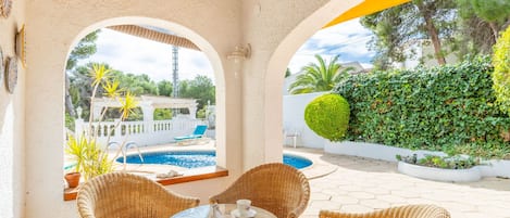 Un área de estar exterior techada con mobiliario de mimbre y una mesa de vidrio, con vistas a una piscina y exuberante vegetación.