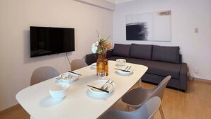 Una sala moderna con una mesa de comedor blanca para dos personas, un sofá gris y un televisor de pantalla plana montado en la pared.