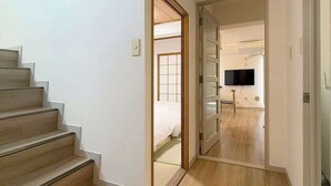 A modern interior with a wooden staircase, a hallway leading to a bedroom with a bed and a TV, and a bathroom with a toilet and sink. - Stylish 2-Story Apartment Near Atomic Bomb Dome (Hiroshima)