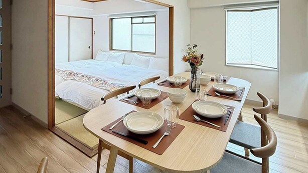 A room with a bed, a dining table set for two, and a small table with a vase of flowers. - Stylish 2-Story Apartment Near Atomic Bomb Dome (Hiroshima)