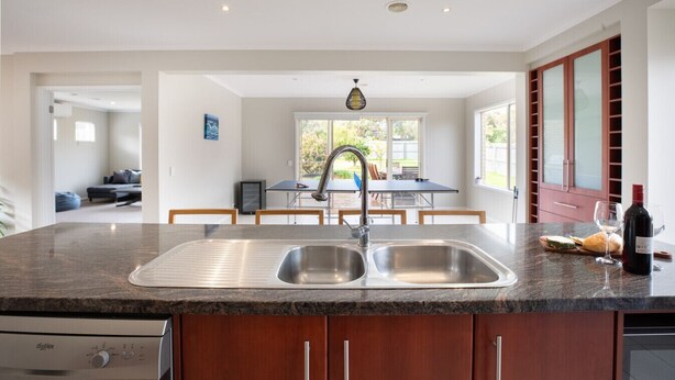 A modern kitchen with a granite countertop, stainless steel sink, and wooden cabinets. There is a dining area with a table and chairs visible through an open door. - Daintree Family Getaway- Beach, Aircon, Great yard (Ocean Grove)