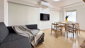 A modern living room with a grey sofa, a dining table with chairs, a flat-screen TV mounted on the wall, and a ceiling light. - Quiet 3BR Near Downtown Kitchen & Dining Equipped (Hiroshima)