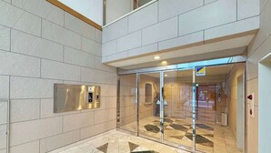Een moderne lobby met een vloer met een geometrisch patroon, een glazen ingang en een aan de muur bevestigde bedieningspaneel voor de lift.