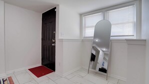 Een nette, witte kamer met een donkere deur, een rode deurmat en een raam met zonwering.
