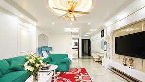 A modern living room with a green sofa, a white coffee table with a vase of flowers, a red patterned rug, a ceiling fan, and a large flat-screen TV mounted on the wall. - Legacy Hotel Ninh Binh (Hoa Lu)