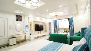 A modern hotel room with a large bed, green upholstered seating, a flat-screen TV, a vanity with a mirror, and a balcony with blue curtains. - Legacy Hotel Ninh Binh (Hoa Lu)