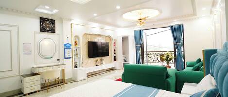 Una habitación de hotel moderna con una cama grande, asientos tapizados en verde, un televisor de pantalla plana, una tocador con espejo y un balcón con cortinas azules.