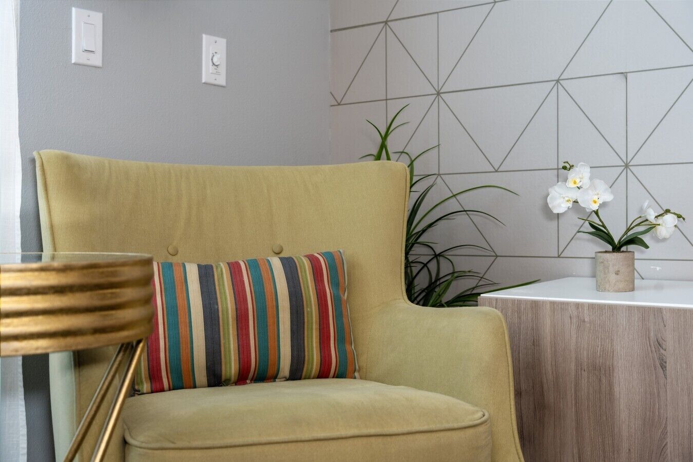 A room with a large armchair, a striped pillow, a small table with a plant, and a geometric wall design.