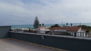 Un balcon avec un sol carrelé et une balustrade. Au fond, on aperçoit des bâtiments aux toits recouverts de tuiles rouges et une vue sur la mer.