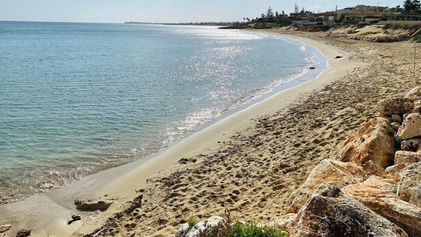 Une plage avec une eau claire, un rivage rocheux et des bâtiments en arrière-plan.