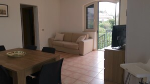 Un salon avec un canapé beige, une table à manger en bois avec des chaises, un téléviseur posé sur un meuble et un balcon donnant sur un paysage verdoyant.