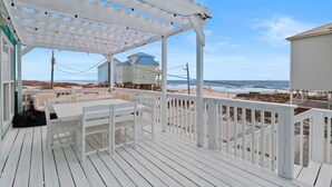 Eine überdachte Terrasse mit Tisch und Stühlen, mit Blick auf einen Strand und Gebäude im Hintergrund.
